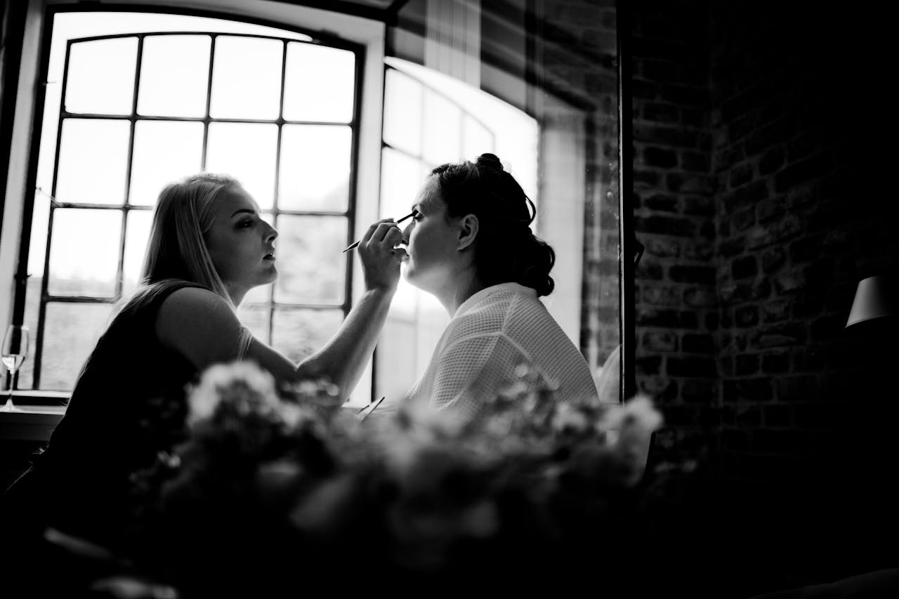 A stylist applies makeup to a woman by a window, captured in elegant black and white.