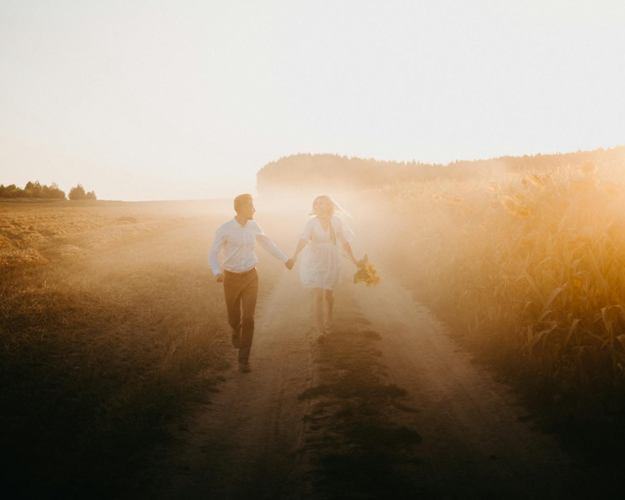 A couple holding hands and walking along a sunlit dirt road during sunset in a rural field.