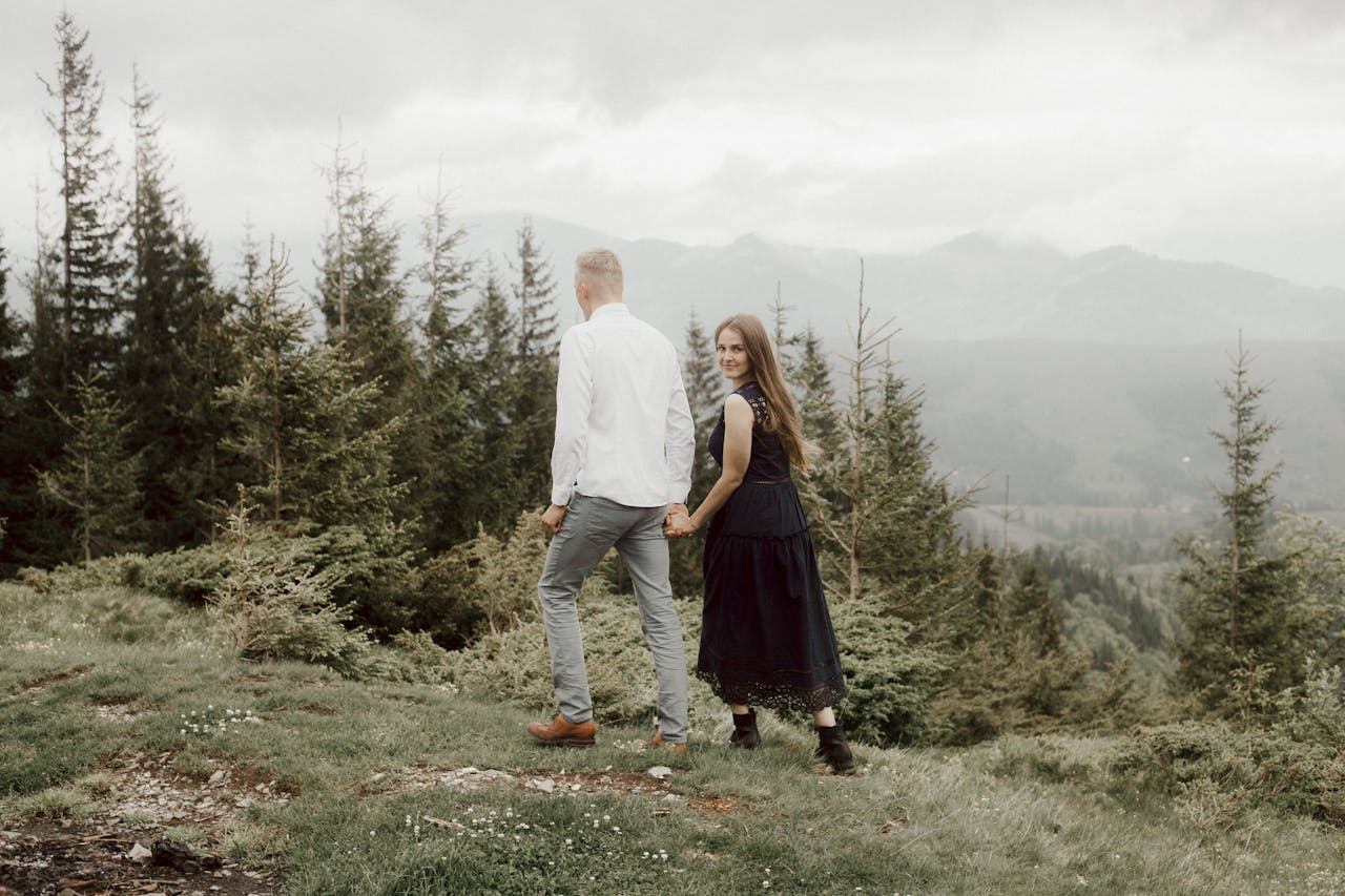 A couple walks hand in hand through a lush mountain meadow, surrounded by tall pine trees.
