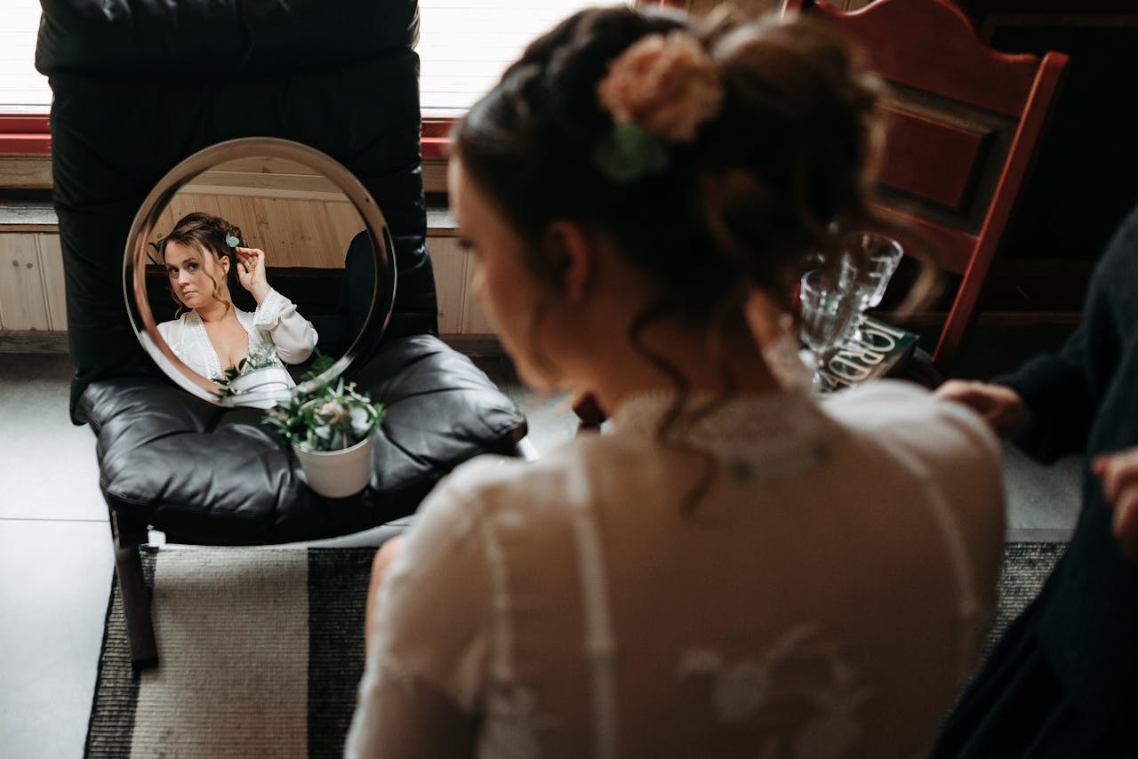 A bride adjusts her hair while looking into a round mirror indoors.